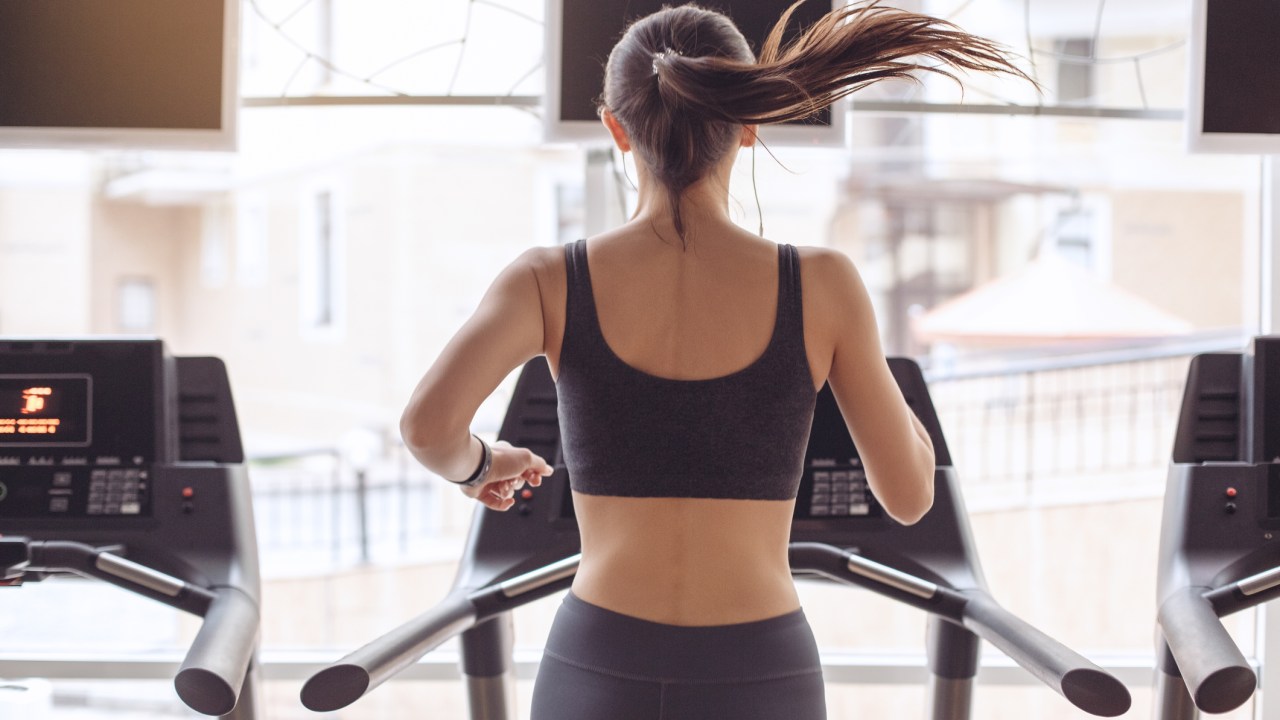 Mulher correndo na esteira fotografada de costas - Como fazer um aquecimento eficaz antes do treino - correr na esteira do prédio ou da academia