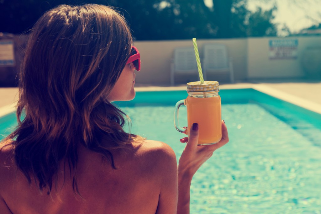 Mulher de costas à beira da piscina segurando um suco