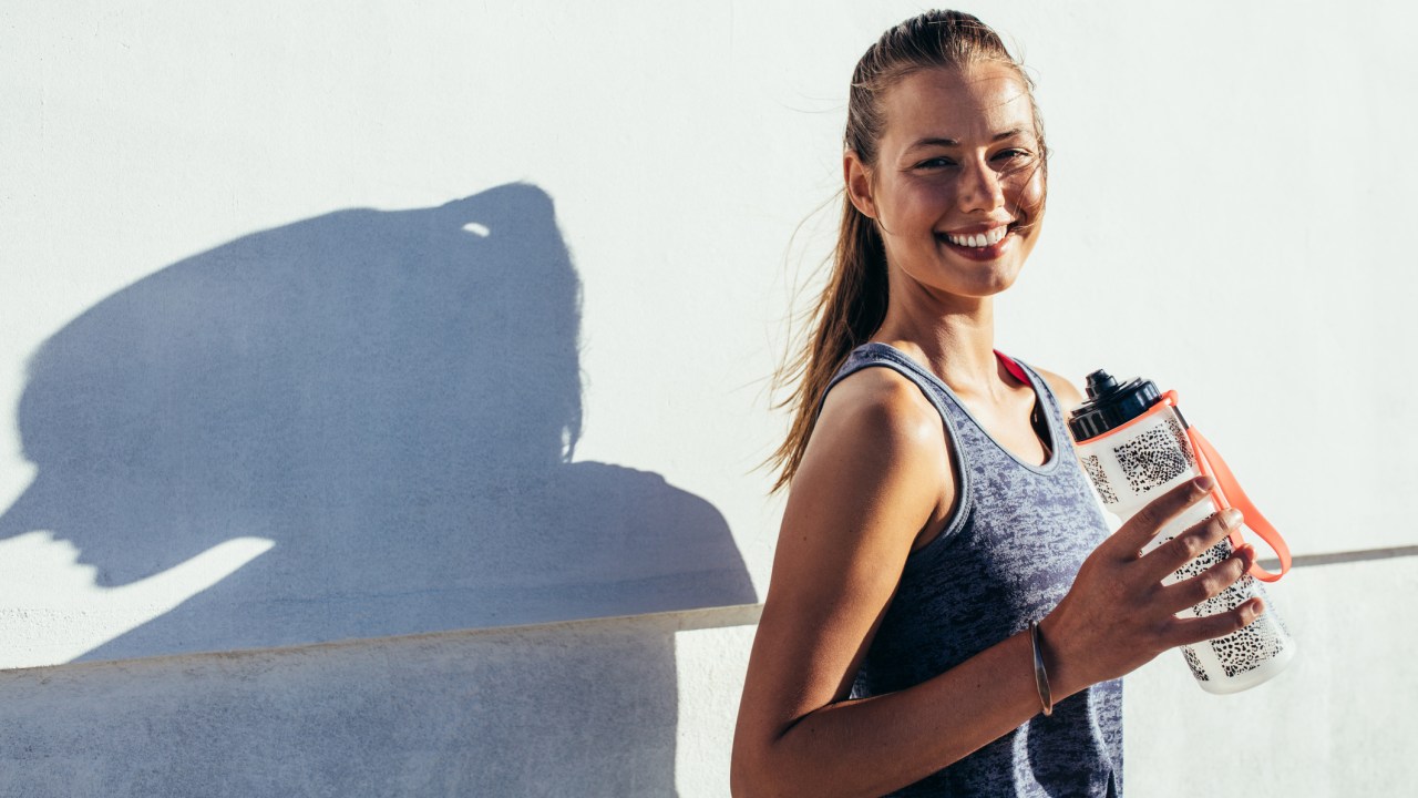 Mulher com roupa fitness fotografada numa parede clara segurando a garrafinha de água - alimentos para evitar cãibra