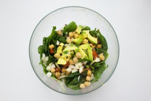 Uma salada de folhas verdes com grão de bico, pedaços de abacate, cebola e tomate em um recipiente transparente em um fundo branco