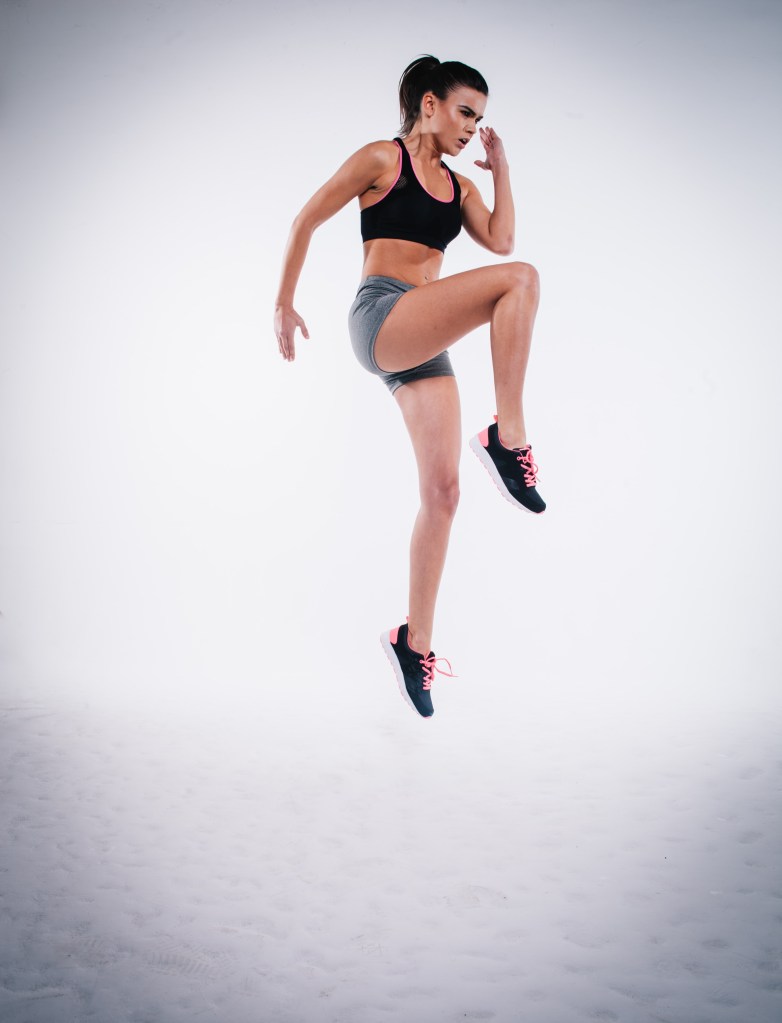 mulher saltando_exercícios dinâmicos Imagem ilustrativa que simula um exercícios chamado Hopserlauf, em que o joelho deve ser elevado até mais ou menos a linha do quadril e os braços devem ser elevados de forma intensa aproximadamente no ângulo de noventa graus.