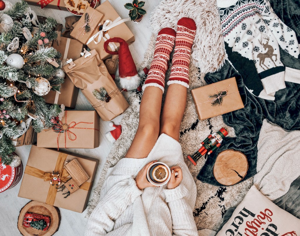 Mulher sentada de pijamas no chão tomando um chocolate quente e cercada de decoração de Natal