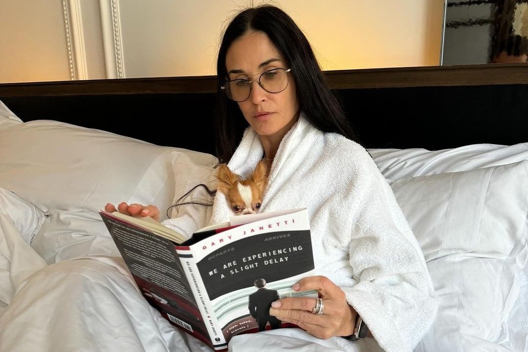 Demi Moore na cama lendo um livro com o seu cãozinho no colo